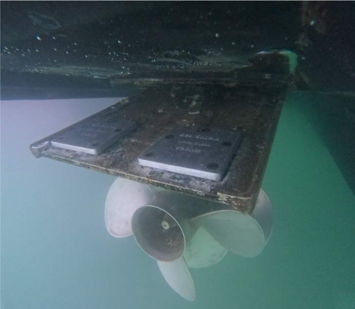 Close-up underwater view of a professionally cleaned boat propeller and metal plates, showing a smooth surface free of algae and "white coral" growth to ensure maximum vessel performance.